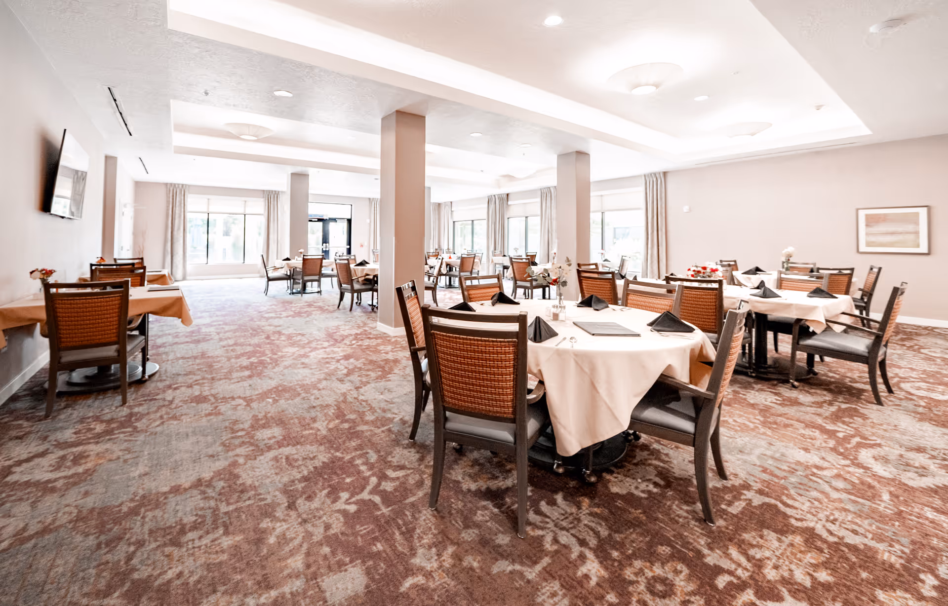 Bright dining room with multiple round and rectangular tables set with white tablecloths and chairs in a senior living facility.