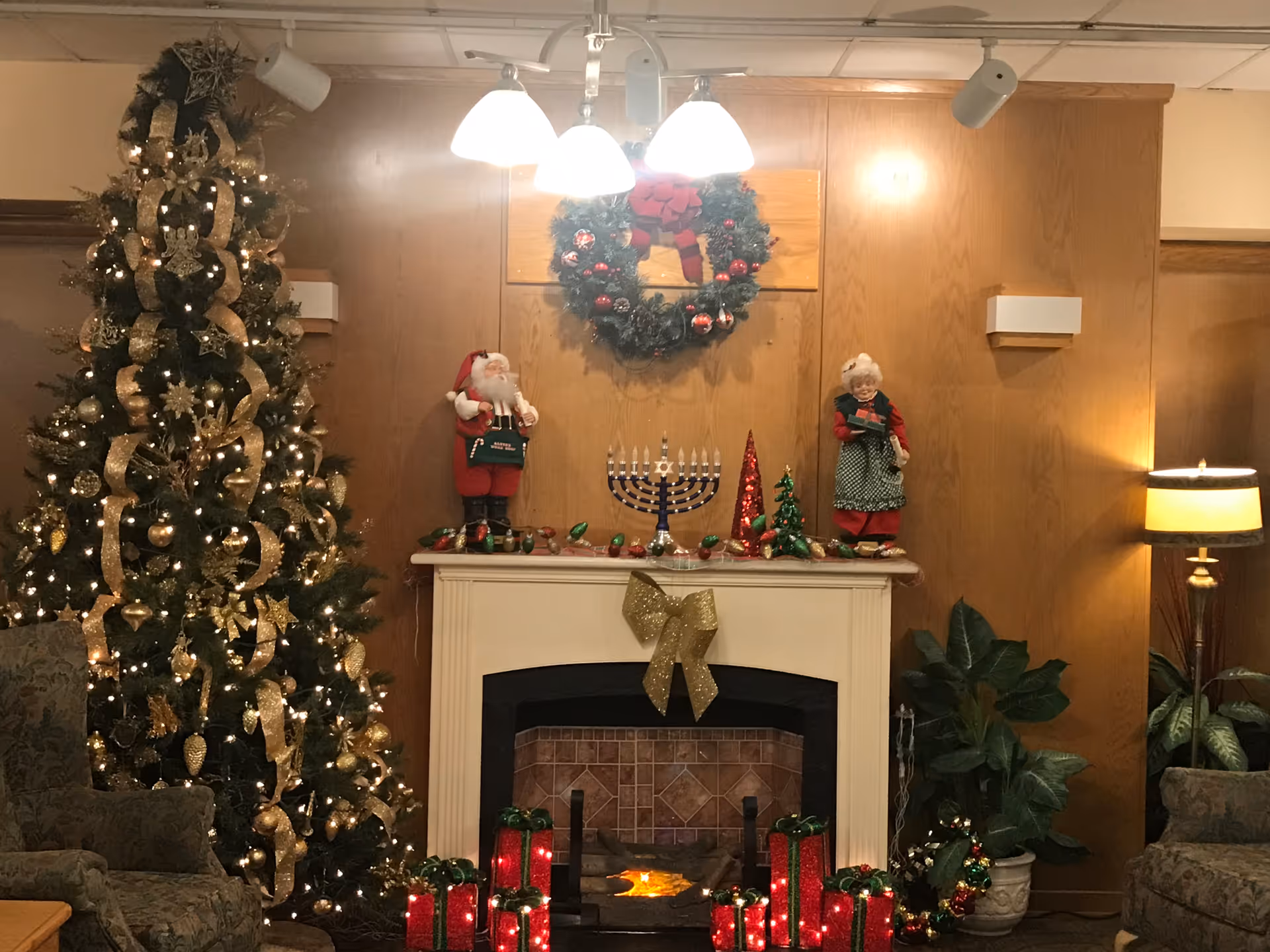 A cozy living room decorated for the holidays with a tall Christmas tree adorned with gold ribbons and ornaments on the left, a white fireplace in the center with a gold bow, a menorah, holiday figurines of Santa Claus and Mrs. Claus, and a wreath hanging above. There are also wrapped gift decorations and a floor lamp on the right side next to green plants and upholstered chairs.