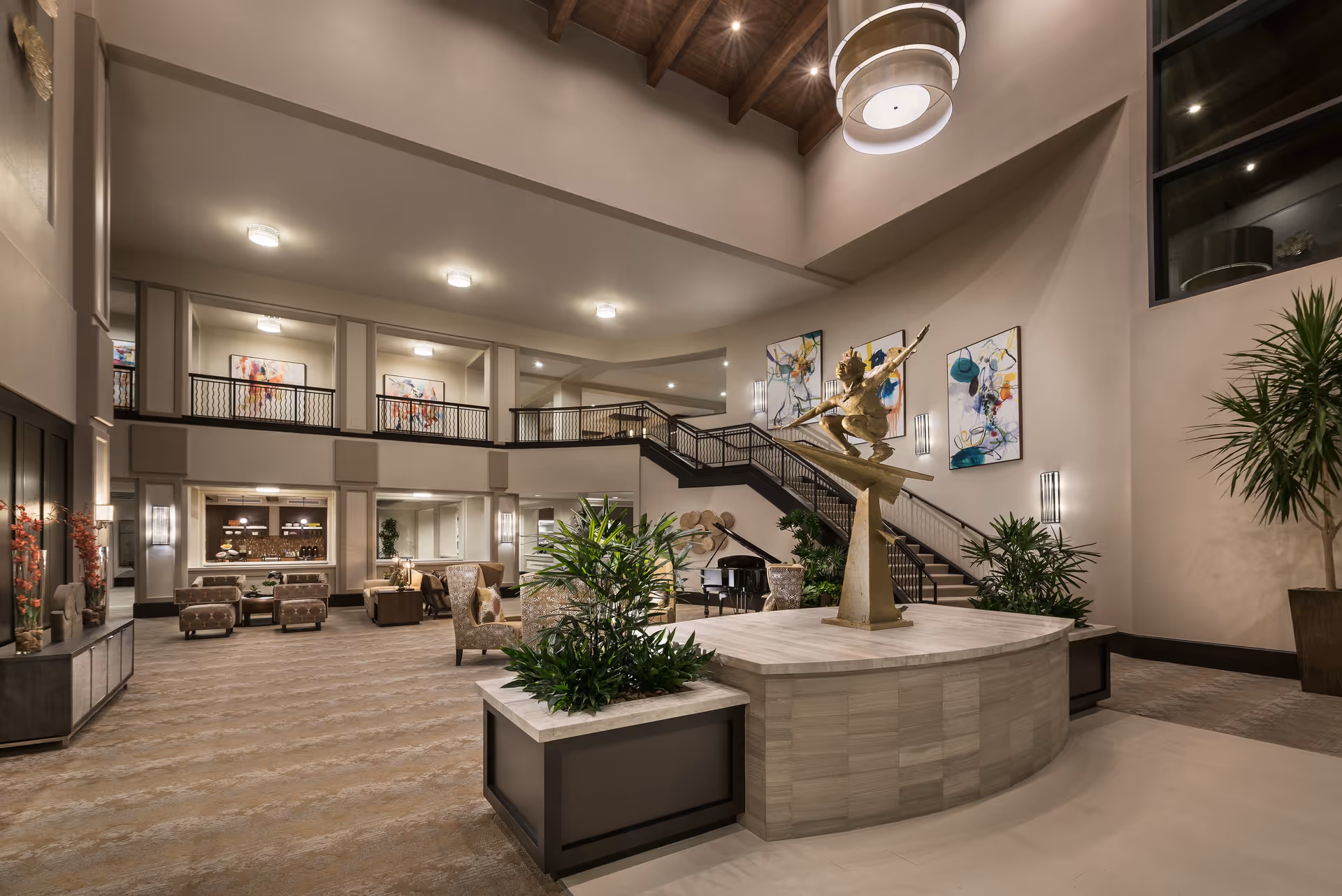 Spacious and well-lit senior living facility lobby with a large sculpture of a figure holding a spear on a raised platform surrounded by plants. The area features comfortable seating, a grand piano, modern artwork on the walls, and a staircase leading to an upper balcony with railings. The ceiling is high with exposed wooden beams and a large circular light fixture.