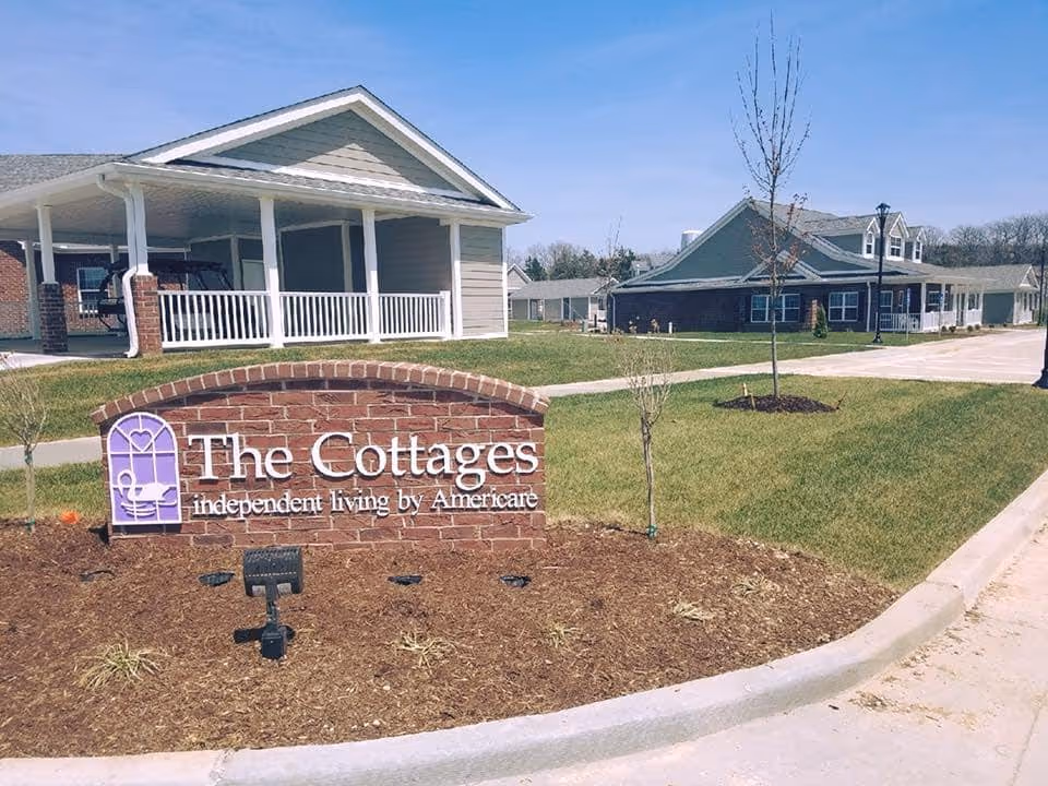 Brick entrance sign reading "The Cottages independent living by Americare" in front of single-story cottage-style senior living buildings and lawns.