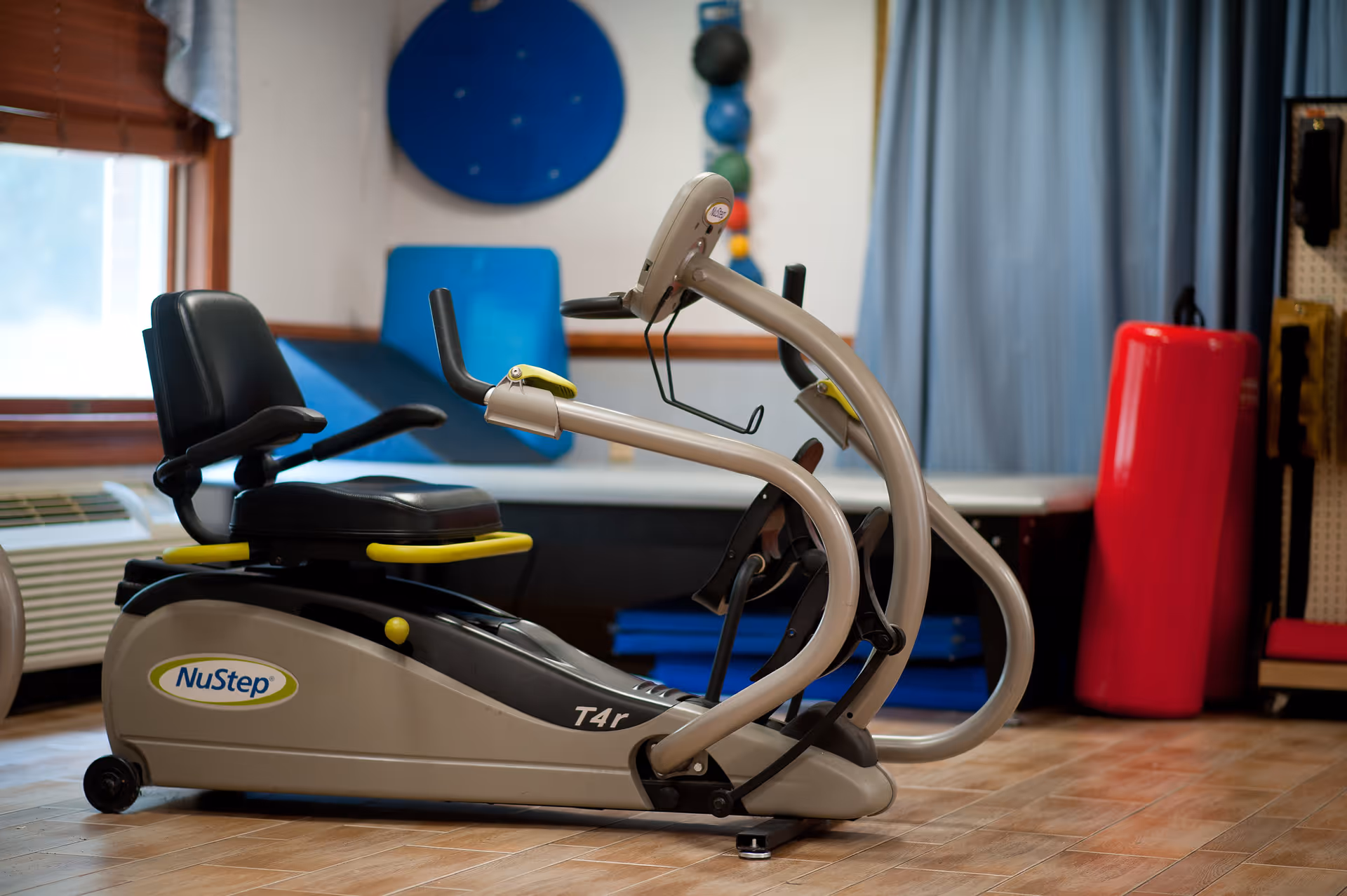 A NuStep recumbent exercise machine in a therapy/fitness room with mats and equipment in the background.