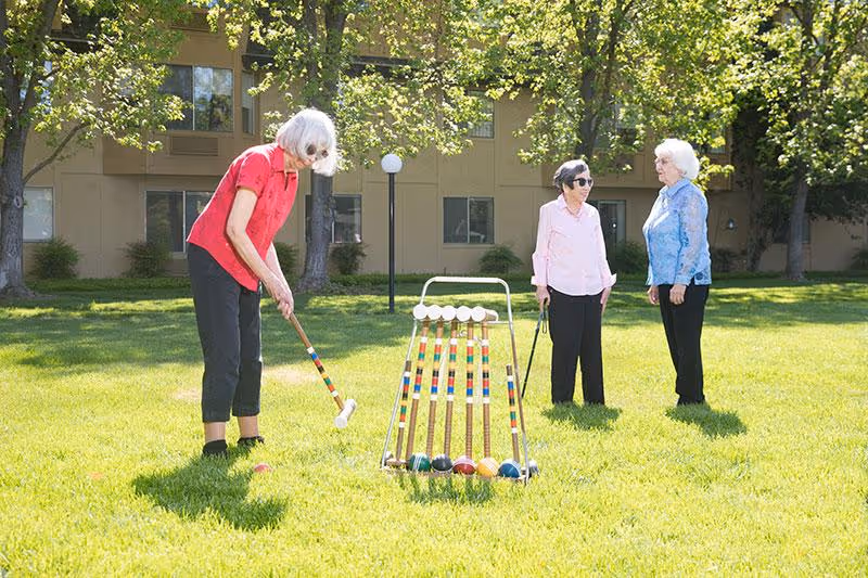 Three elderly women outdoors on a grassy lawn in front of a building. One woman in a red shirt is playing croquet, preparing to hit a ball with a mallet. The other two women, one in a pink shirt and the other in a blue shirt, are standing nearby watching and talking. Trees and windows of the building are visible in the background.