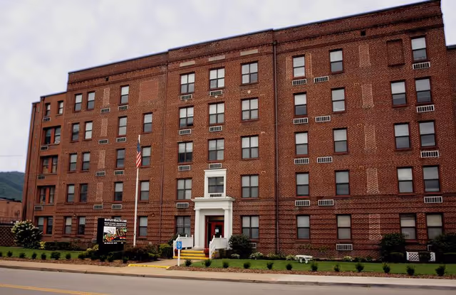 Front exterior view of a large, multi-story brick building with multiple windows and a white entrance with steps. There is an American flag on a flagpole near the entrance and a sign that reads 'Maples Health Care'. The building is surrounded by a small lawn and bushes.
