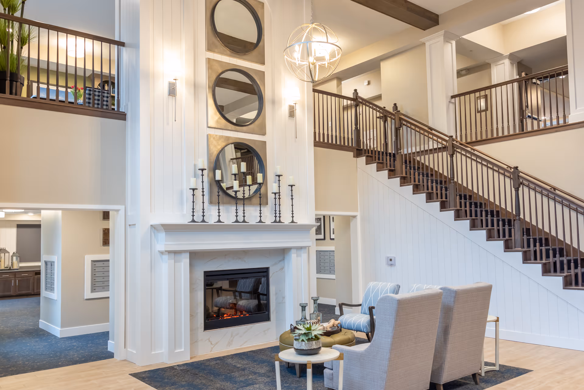 A bright and spacious senior living facility common area with a modern fireplace, three round mirrors mounted vertically above it, and tall candle holders on the mantel. There are two upholstered armchairs and a small round table with decorative items in front of the fireplace. A staircase with dark wooden railings leads to an upper level with a balcony overlooking the area. The walls are painted in neutral tones, and the space is well-lit with natural and artificial lighting.