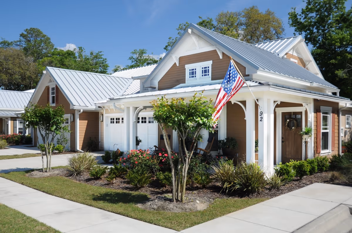 Exterior view of a single-story residential building with a metal roof, beige siding, white trim, and a front porch. The porch has two rocking chairs and an American flag mounted on a pole. There are small trees, bushes, and flowers planted in the landscaped yard around the building. The sky is clear and blue.