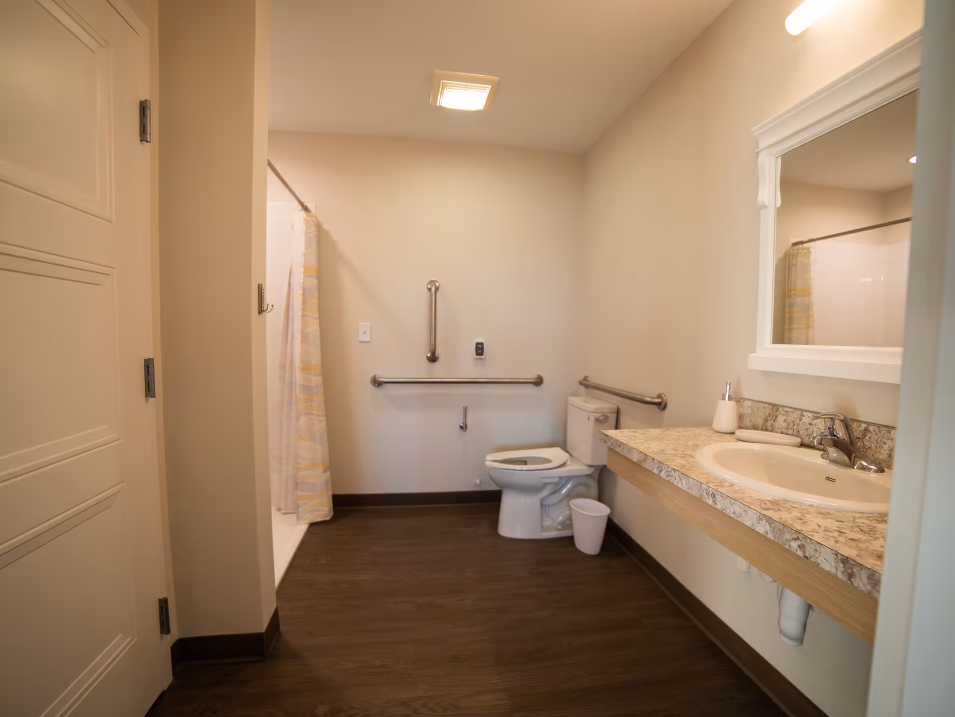 A clean and accessible bathroom with a toilet equipped with grab bars on the walls, a shower with a curtain, a sink with a granite countertop, a mirror above the sink, and a small trash bin on the floor. The bathroom has beige walls and dark wood flooring.
