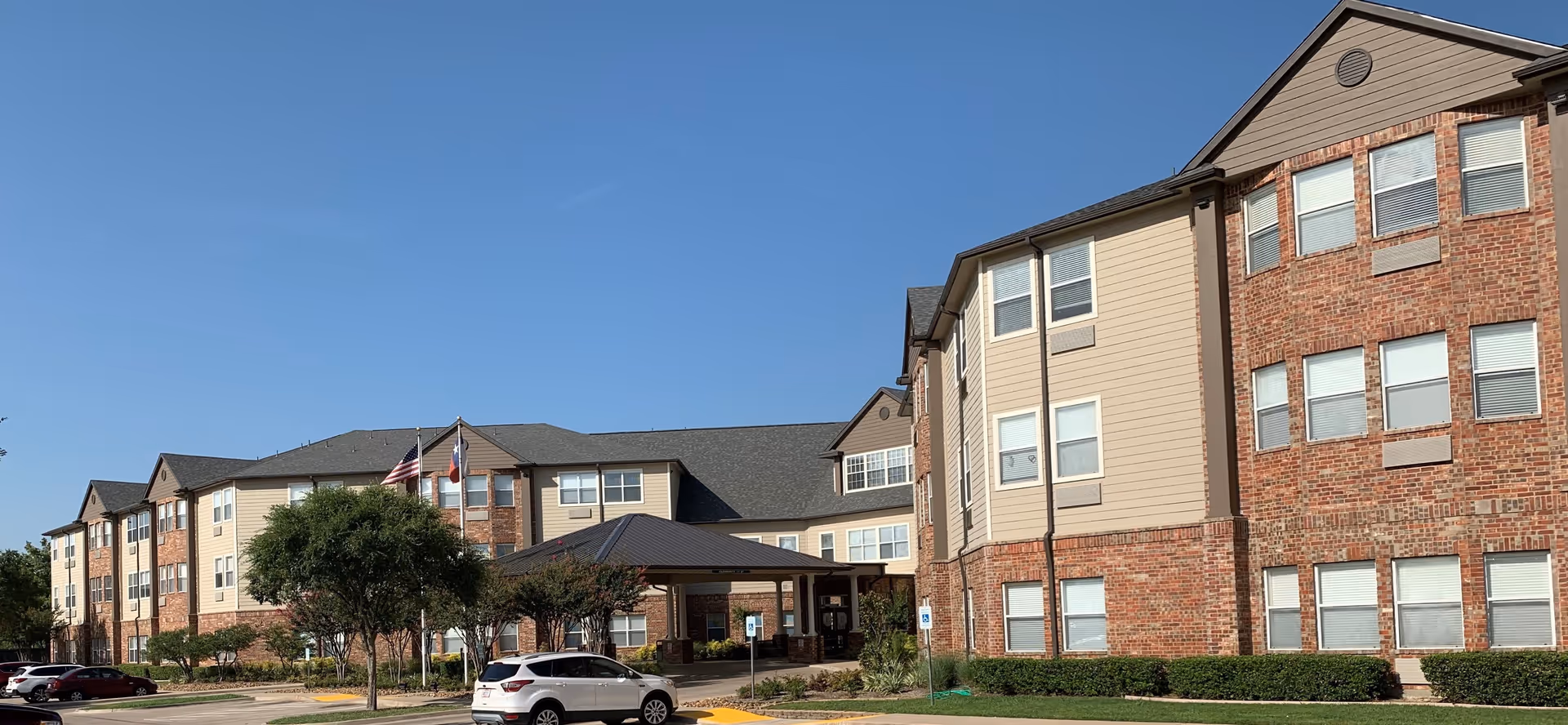Exterior view of Emerson South Collins senior living facility showing a large three-story building with a combination of brick and beige siding, multiple windows, a covered entrance, several parked cars, and two flagpoles with flags against a clear blue sky.