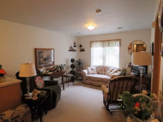 A cozy living room with a floral-patterned sofa, a dark armchair, a wooden rocking chair, and various decorative plants and items. There is a window with sheer curtains letting in natural light, a table lamp, and framed artwork on the wall.