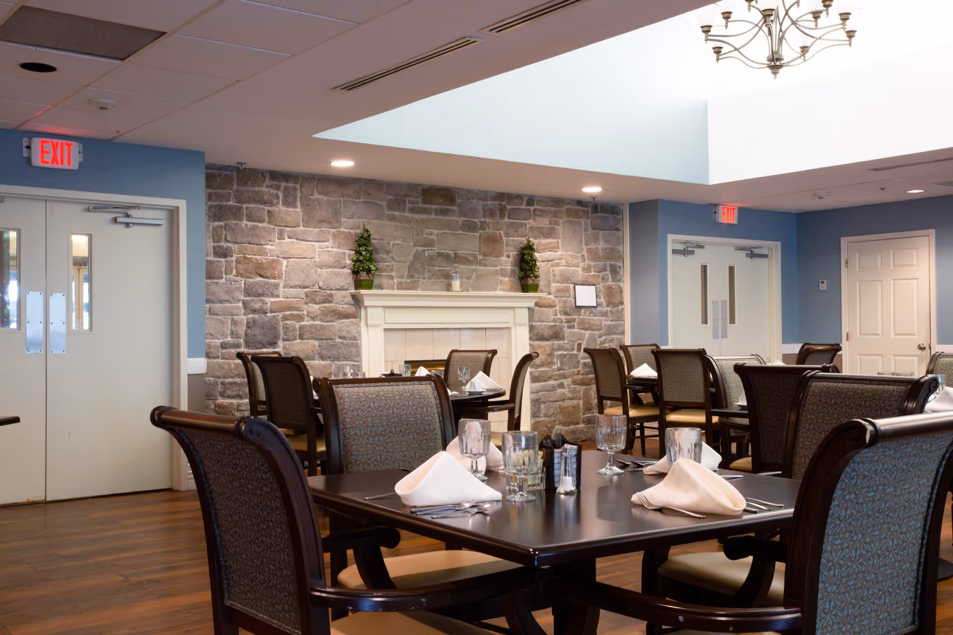 A dining room in Morningstar Assisted Living featuring multiple tables set with white napkins, glasses, and silverware. The room has wooden floors, a stone accent wall with a white mantel decorated with small plants, and blue walls. There are exit signs above double doors on two sides of the room.