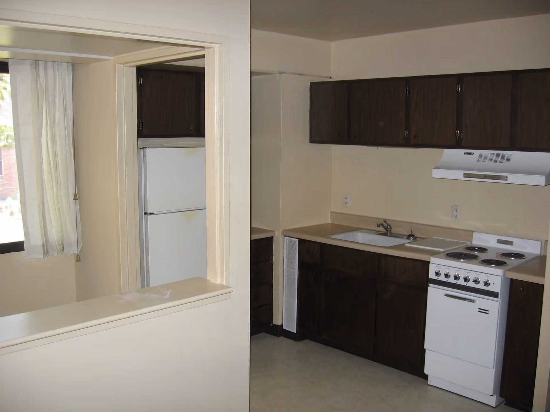Small kitchen area with dark wooden cabinets, a white electric stove with four burners, a white range hood, a sink with a faucet, and a white refrigerator partially visible through a pass-through window with cream-colored walls and a window with white curtains.