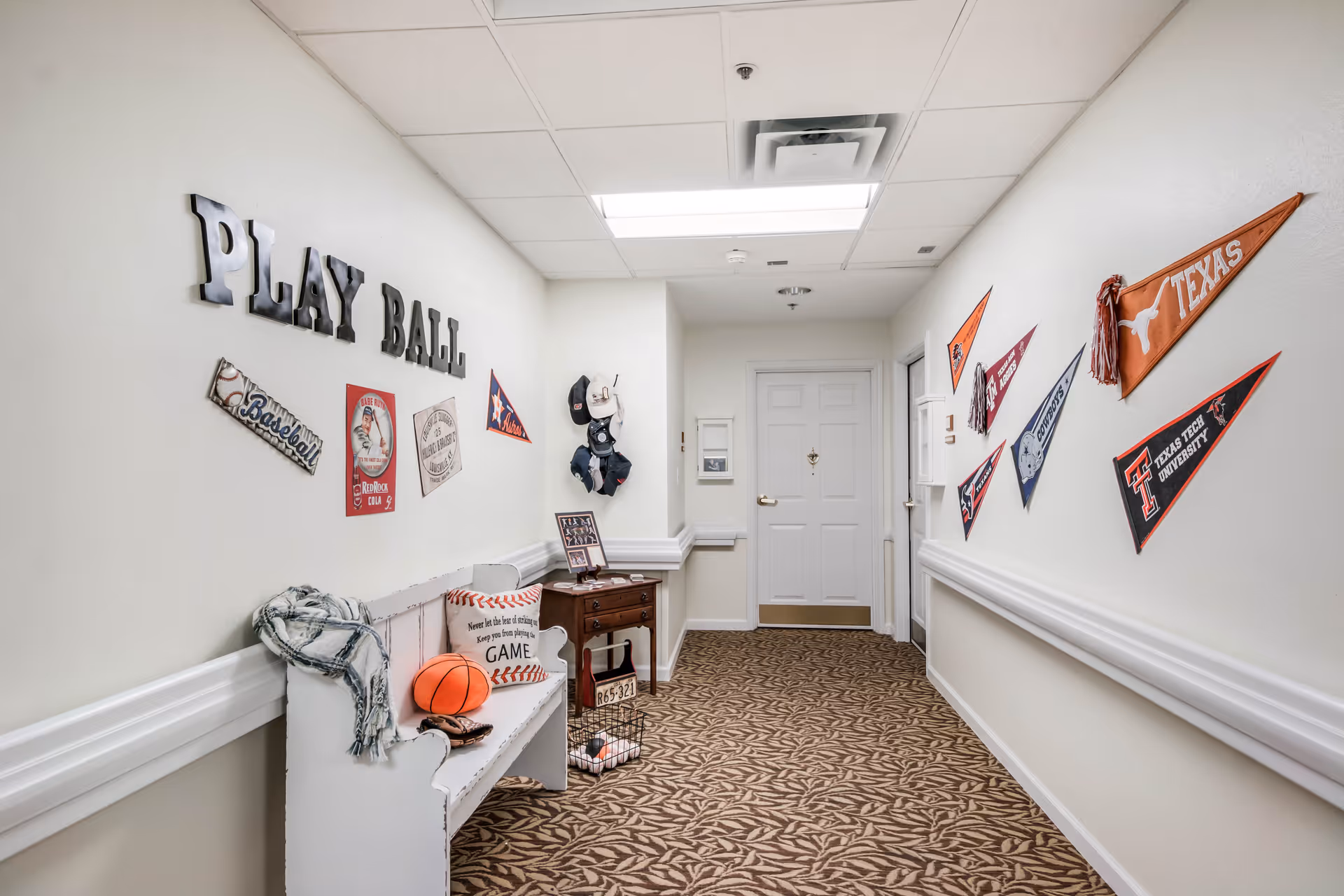 A hallway decorated with sports-themed items including a white bench with a basketball, a baseball glove, and a pillow. The wall on the left has metal letters spelling 'PLAY BALL' along with baseball-themed signs. The wall on the right displays several college pennants. The floor is carpeted with a leaf pattern, and there is a white door at the end of the hallway.