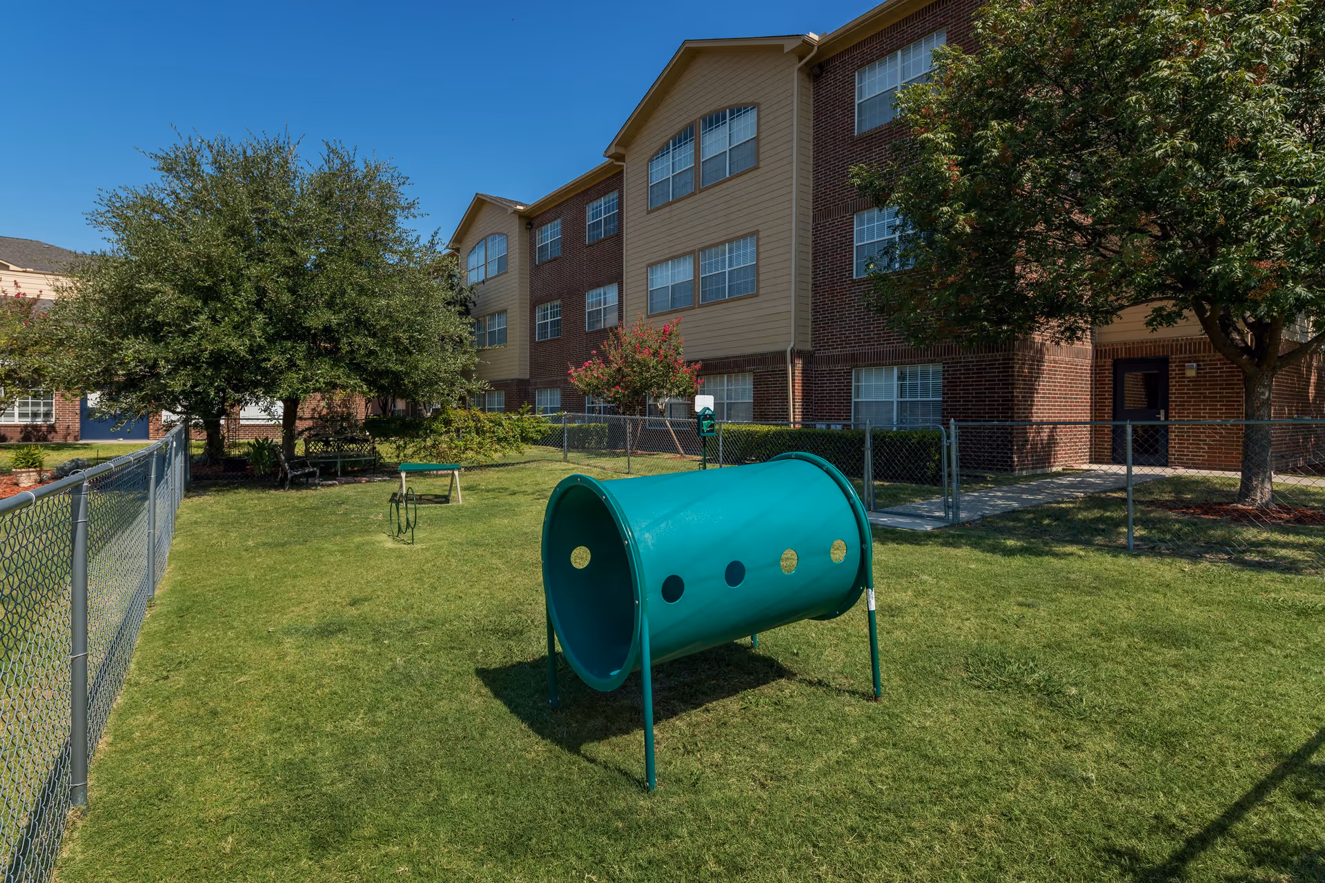 Outdoor fenced grassy area with a green cylindrical tunnel play structure, trees, and a three-story brick and beige building in the background under a clear blue sky.