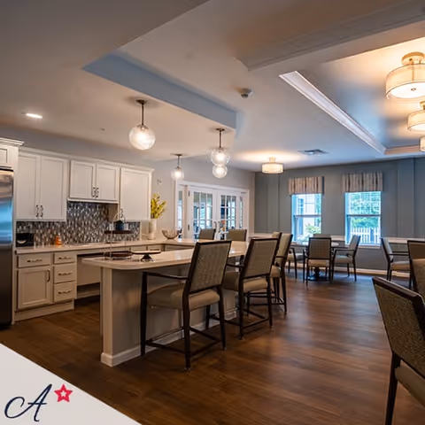 A bright and spacious kitchen and dining area in an assisted living facility, featuring white cabinetry, a kitchen island with bar stools, pendant lighting, and a dining table near windows with natural light.