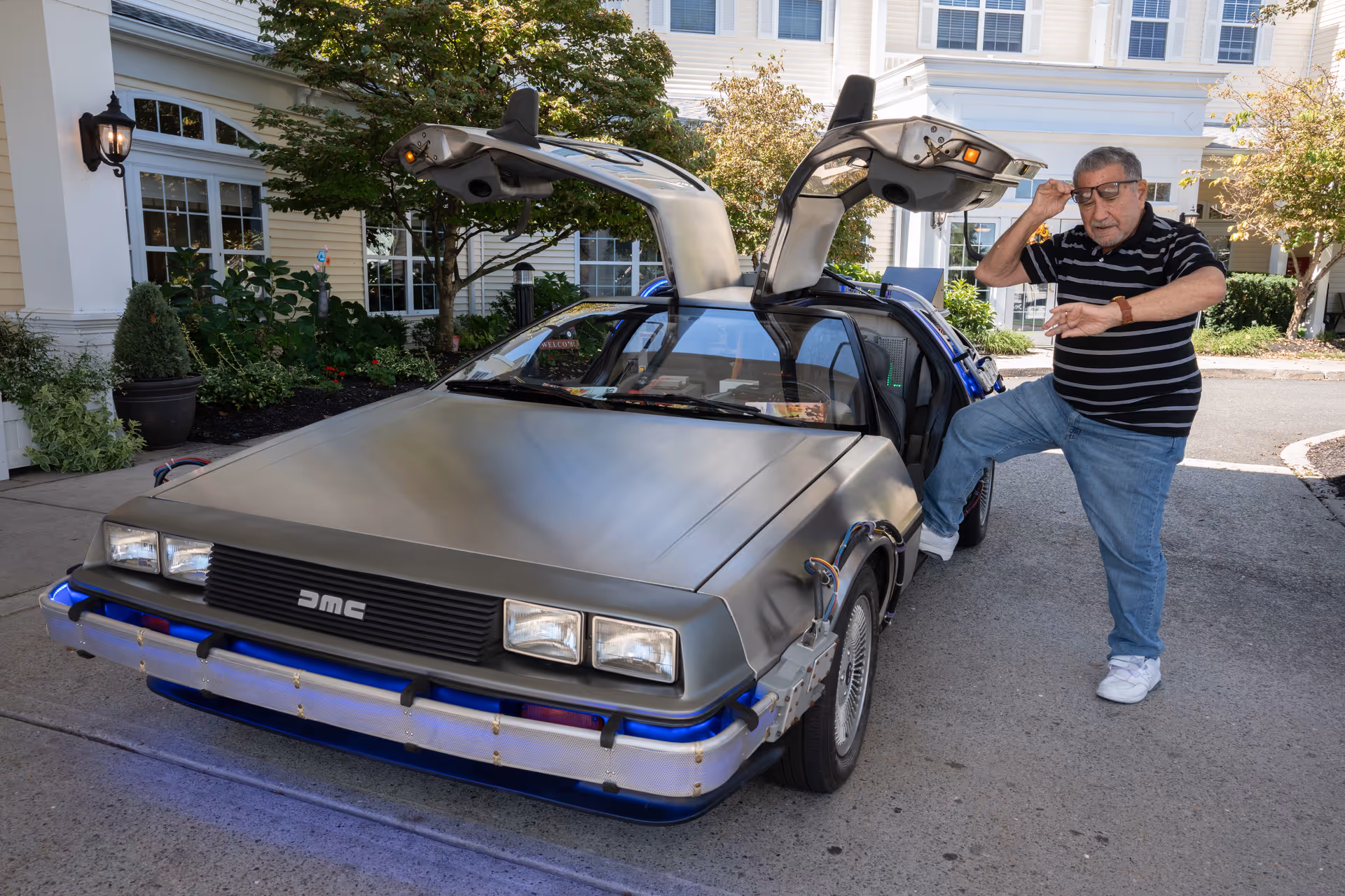 A DeLorean with its gull-wing doors open is parked by a senior living facility entrance while an older man poses beside it.