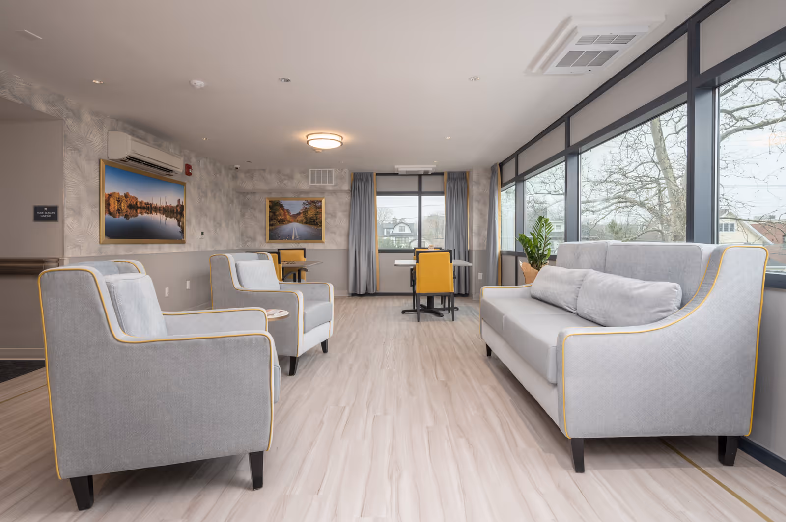 Bright senior living lounge with gray sofas and armchairs arranged facing large windows.