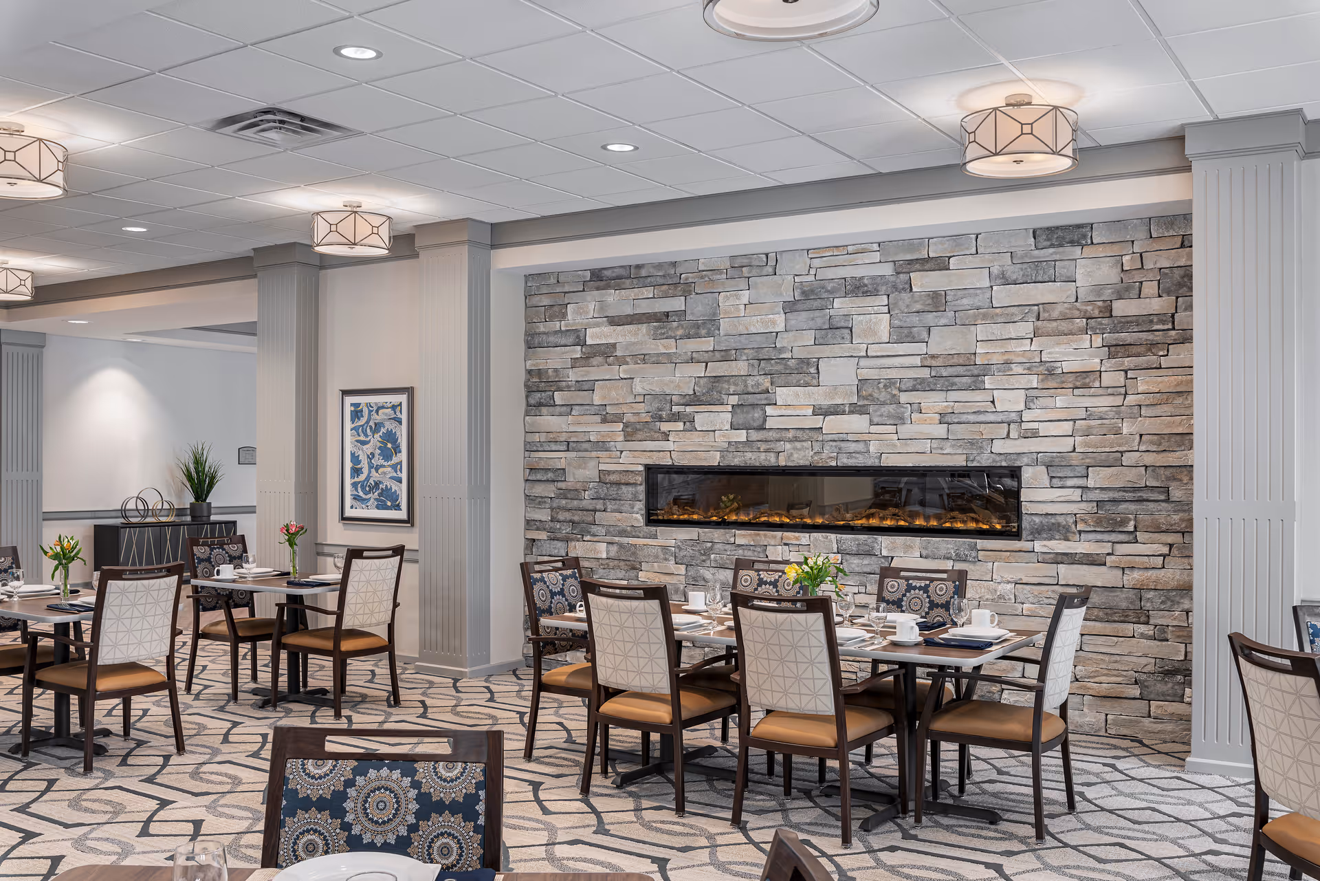 Dining room with multiple set tables and chairs arranged in front of a stone accent wall featuring a long linear fireplace.