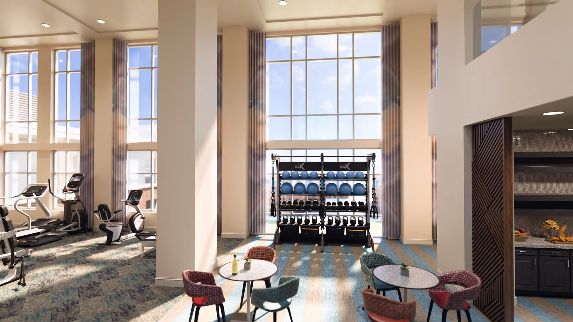 A bright and spacious fitness area with large floor-to-ceiling windows letting in natural light. The room features exercise equipment including treadmills and stationary bikes on the left side, a rack with various exercise balls and weights in the center, and two small round tables with colorful chairs in the foreground. To the right, there is a partial view of a kitchenette or refreshment area with a countertop and fruit bowl.