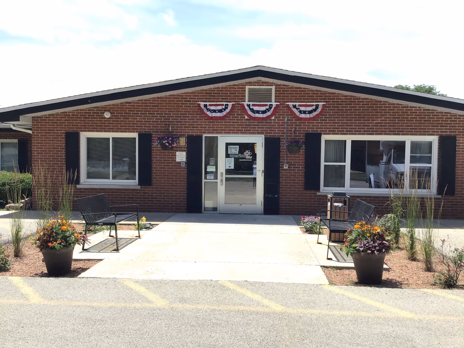 Front exterior view of a single-story brick building with a peaked roof. The entrance has glass double doors flanked by black shutters and two windows. There are three patriotic red, white, and blue bunting decorations above the door. Two black metal benches and large flower pots with colorful flowers are placed on either side of the entrance walkway.