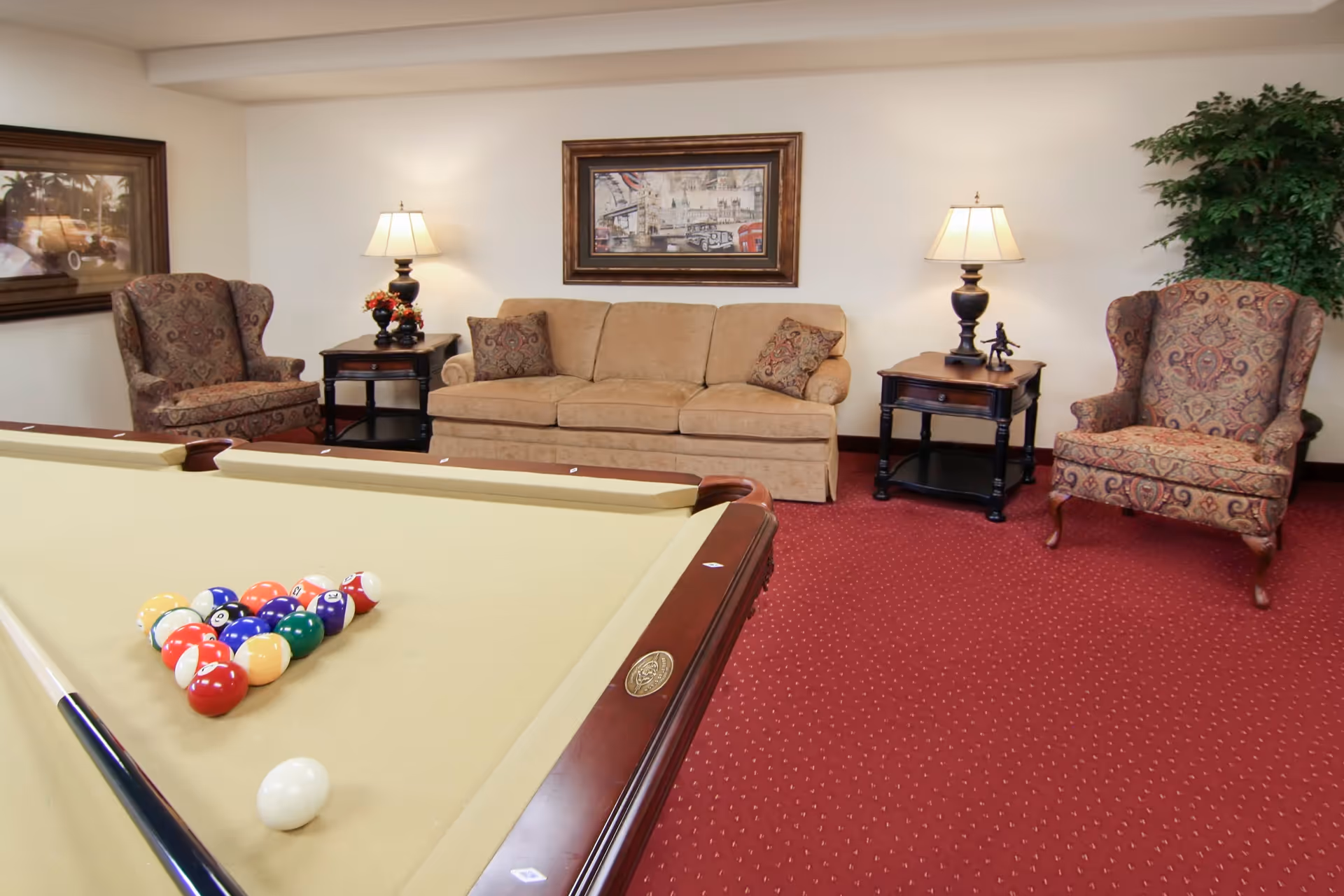 A cozy living room area featuring a beige sofa with two patterned cushions, two matching patterned armchairs, two side tables each with a lamp, a framed picture on the wall, a large green plant in the corner, and a pool table with billiard balls arranged in a triangle and a cue stick resting on the table.