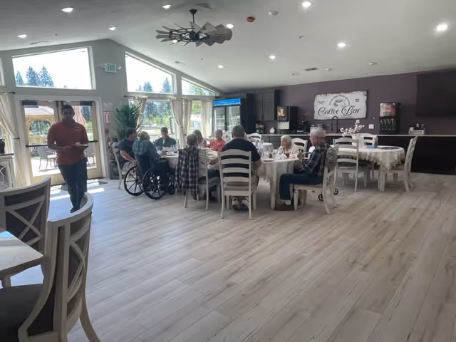 A group of elderly people seated around a table in a bright dining room with large windows and light wood flooring. A staff member stands nearby holding a tray. The room features a coffee bar area with a sign on the wall and several empty tables and chairs.