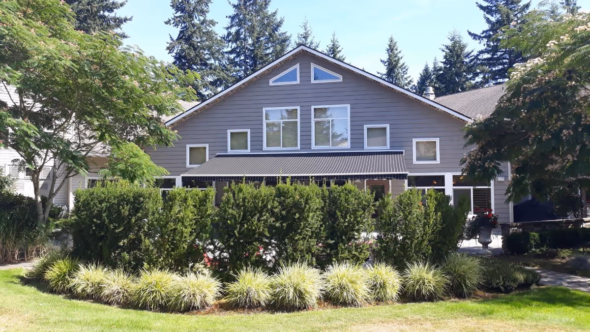 Exterior view of a senior living facility building with gray siding, multiple windows, and a black awning. The building is surrounded by green bushes, ornamental grasses, and trees under a clear blue sky.