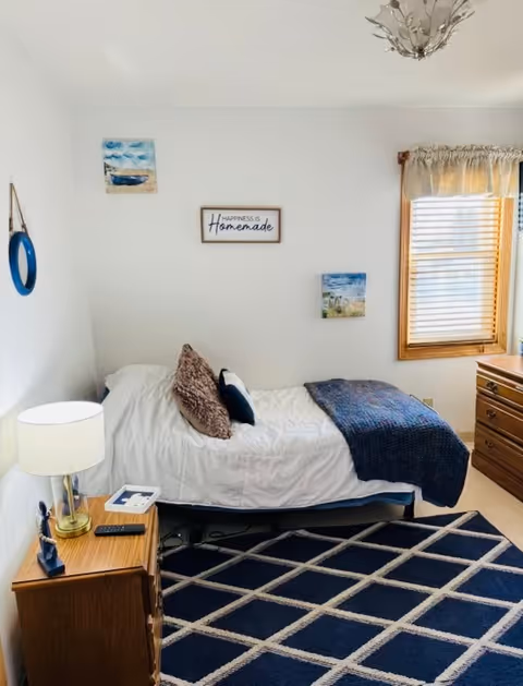 A cozy bedroom with a single bed against a white wall. The bed has white bedding, a dark blue throw blanket, and several pillows. Above the bed, there are three wall decorations including a sign that reads 'Happiness is Homemade'. A wooden nightstand with a lamp, remote control, and small items is next to the bed. A window with wooden blinds and a valance is on the right side, and a wooden dresser is partially visible. The floor has a dark blue rug with a white diamond pattern.