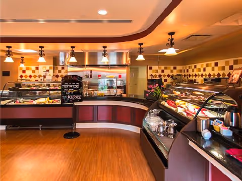 Interior view of a dining area with a curved buffet counter displaying various food items. The room features warm lighting with hanging lamps, a tiled wall with a diamond pattern, and wooden flooring. There is a small chalkboard sign on the counter with a motivational message.