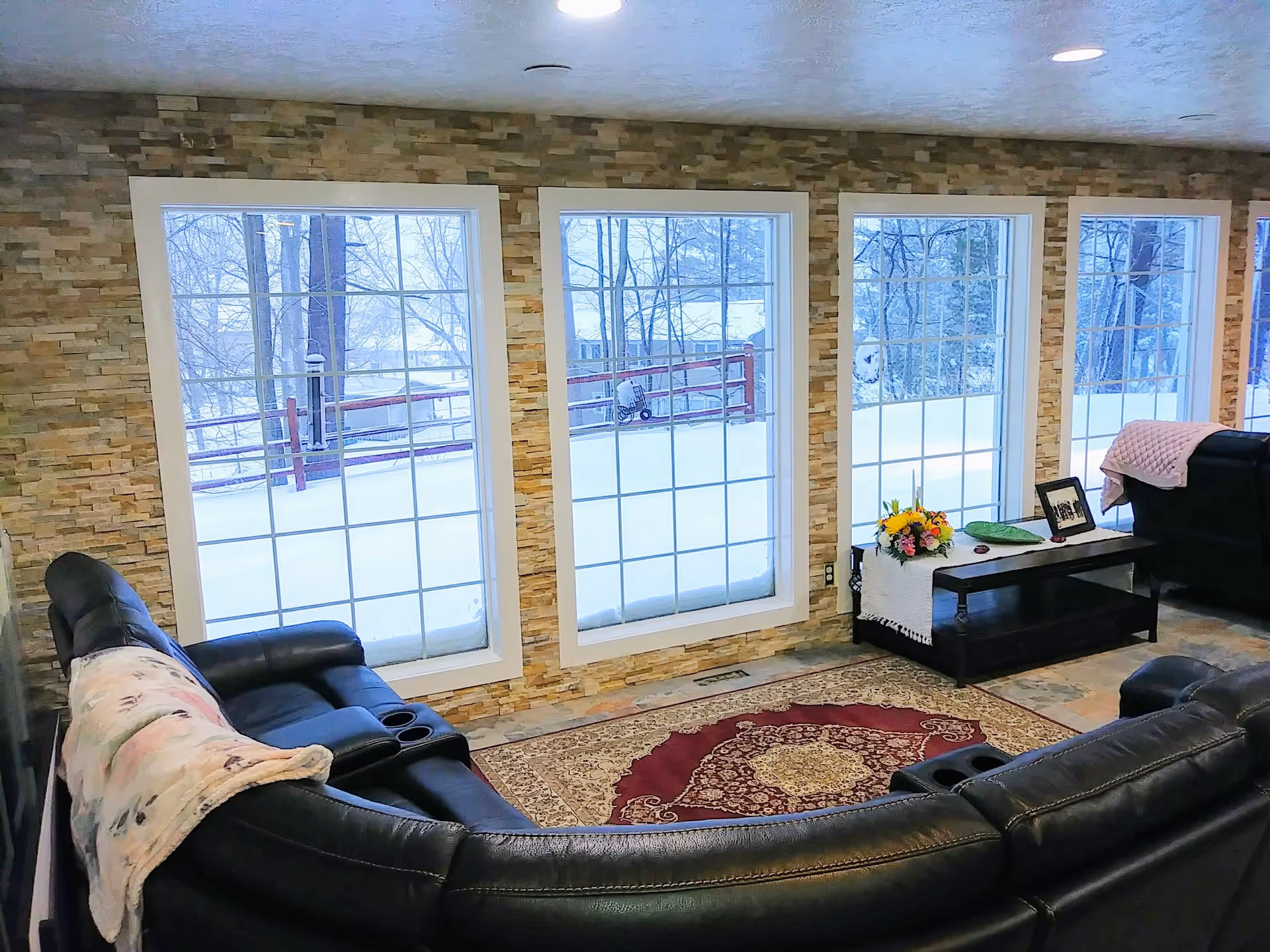 A cozy living room with a large black leather sectional sofa arranged around a patterned area rug. The room features a stone accent wall with four large windows showing a snowy outdoor scene. A dark wooden coffee table holds a bouquet of flowers, a framed photo, and a green decorative plate. The room is well-lit with recessed ceiling lights.