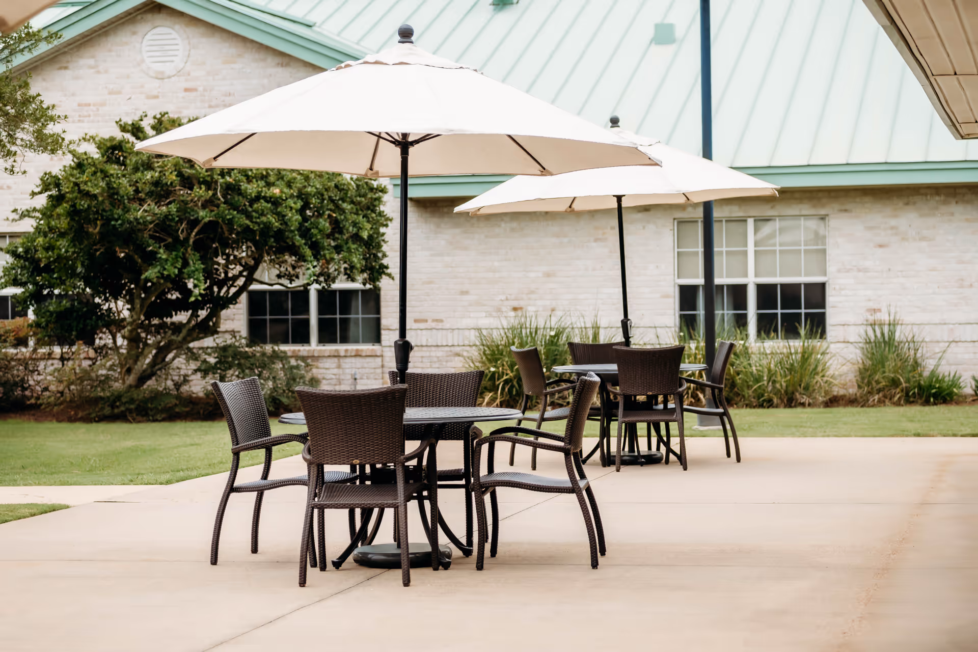 Outdoor patio with wicker tables and chairs under large umbrellas in front of a light-brick building.