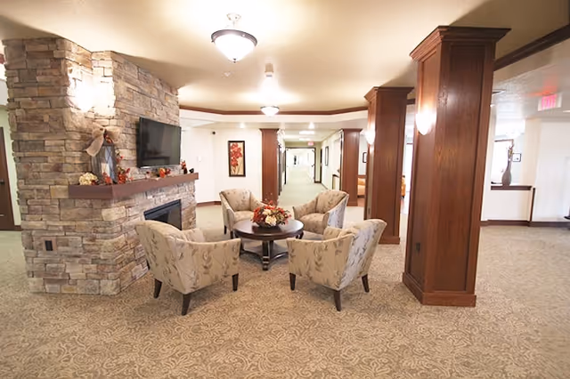 A cozy seating area in a senior living facility with four upholstered armchairs arranged around a round wooden table decorated with a floral centerpiece. The area features a stone fireplace with a mounted flat-screen TV above it. The space is well-lit with ceiling lights and has carpeted floors and wooden columns. A hallway extends in the background with framed artwork on the walls.