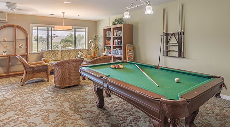 A cozy recreational room with a green felt pool table in the foreground, a cue stick resting on the table, and a rack of pool cues mounted on the wall. In the background, there is a seating area with wicker chairs and a floral-patterned sofa near a large window letting in natural light. A wooden bookshelf filled with books and games is also visible, along with a patterned carpet covering the floor.