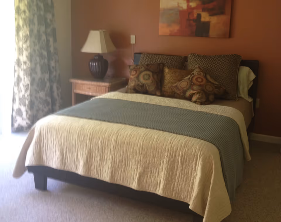 A neatly made bed with a beige quilt and a blue throw blanket in a bedroom. The bed has multiple decorative pillows with various patterns. Next to the bed is a wooden nightstand with a dark lamp. A painting hangs on the wall above the bed, and floral curtains cover a window or door on the left side.