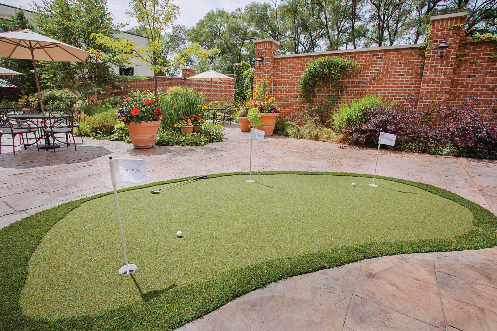 Outdoor putting green with three golf holes and flags, surrounded by a stone patio with potted plants, flowers, and patio tables with umbrellas. A brick wall with greenery is in the background.