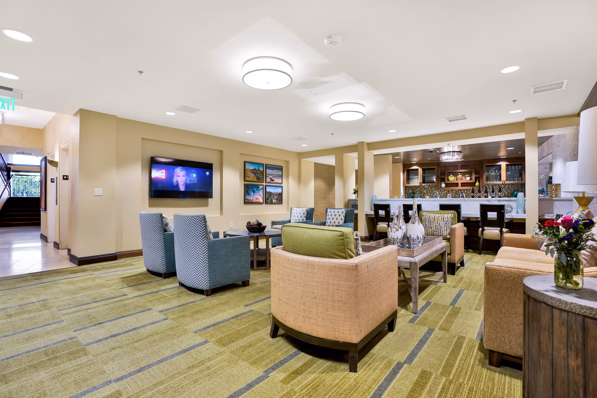 A spacious, well-lit common area in a senior living facility featuring comfortable seating with armchairs and sofas arranged around coffee tables. A flat-screen TV is mounted on the wall, and framed pictures are displayed beside it. In the background, there is a bar area with high chairs and decorative items. The room has a green patterned carpet and neutral-colored walls.