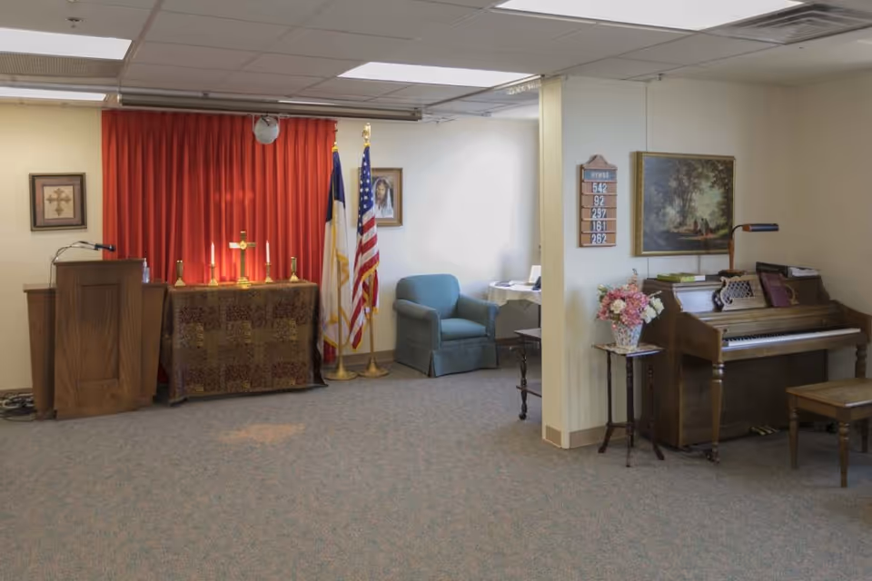 Small chapel/common room with an altar and pulpit on the left, American flags and an armchair in the center, and a piano with a vase of flowers on the right.