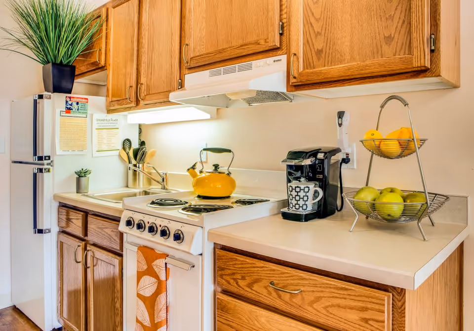 Small kitchen with oak cabinets, a white stove topped by a yellow kettle, a coffee maker, refrigerator, sink, and a two-tier fruit basket.