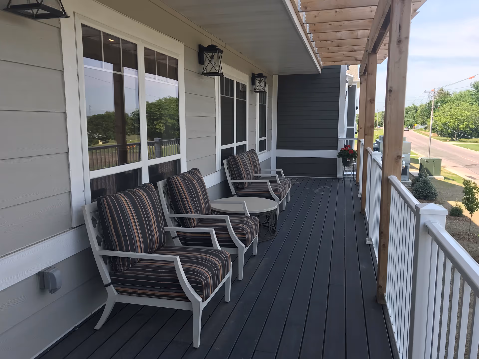 A covered outdoor balcony with three cushioned armchairs arranged around a small round table. The balcony has a dark wooden floor, white railing, and wooden beams supporting the roof. There are large windows on the building wall and a view of a road and greenery in the background.