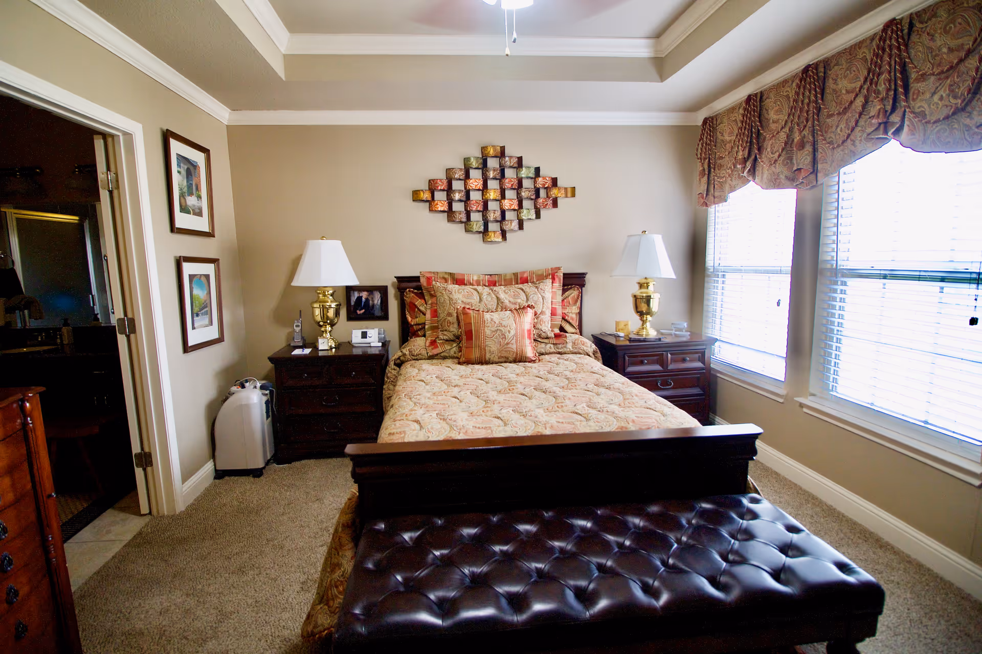 A cozy bedroom with a neatly made bed featuring patterned bedding and multiple pillows. There are two wooden nightstands on either side of the bed, each with a brass lamp. A decorative wall art piece hangs above the bed. To the right, two large windows with blinds and patterned valances allow natural light into the room. A dark leather tufted bench sits at the foot of the bed. On the left side, there is an open door leading to a bathroom.