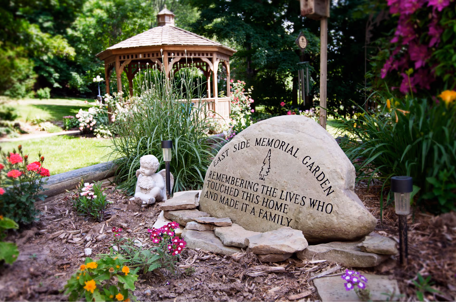 A memorial garden with a large engraved stone that reads 'East Side Memorial Garden Remembering the lives who touched this home and made it a family.' The garden features colorful flowers, a small angel statue, and a wooden gazebo in the background surrounded by lush greenery.