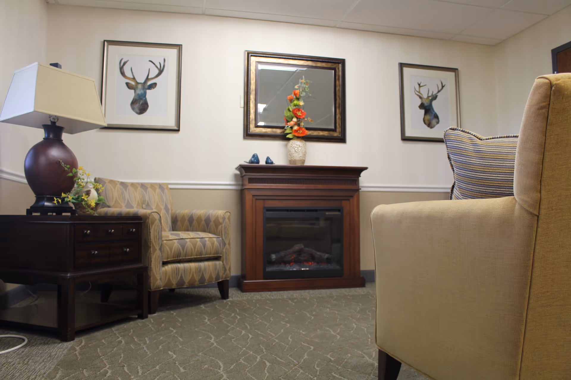 Cozy seating area with armchairs, a side table and lamp, decorative electric fireplace, mirror, and framed deer prints on the wall.