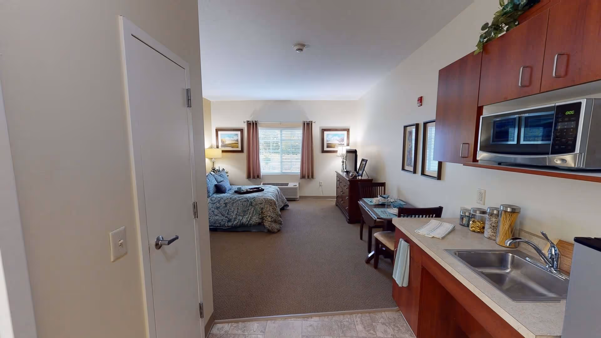 Interior view of a senior living facility studio apartment at The Landing Of Brighton, showing a small kitchen area with a sink, microwave, and countertop on the right, a dining table with two chairs, a dresser with a TV, and a bed with blue patterned bedding near a window with curtains.