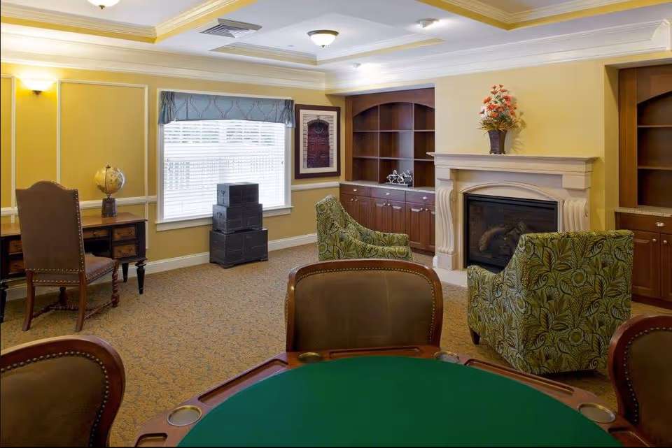 A cozy living room with a green felt poker table in the foreground, two patterned armchairs facing a white fireplace with a floral arrangement on the mantel, built-in wooden shelves and cabinets on either side of the fireplace, a window with blinds and a valance, a wooden desk with a globe on it, and a stack of black boxes near the window.