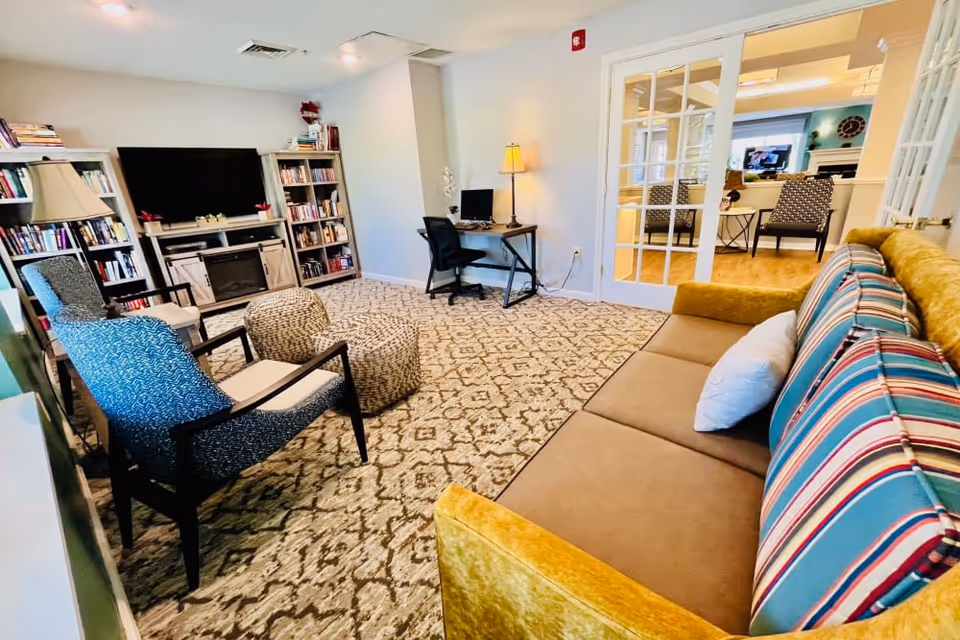 A cozy living room with a patterned carpet, a mustard yellow sofa with striped cushions, two blue armchairs, a round ottoman, a floor lamp, and a desk with a computer. There are bookshelves and a TV with a fireplace below it. French doors open to another room with additional seating.