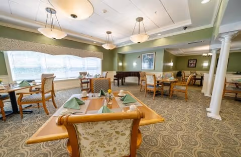 A spacious dining room with several wooden tables and chairs arranged neatly. Each table is set with green folded napkins, plates, and utensils. Large windows with blinds allow natural light to fill the room. The walls are painted green, and there is a grand piano in the corner. Ceiling lights provide additional illumination.
