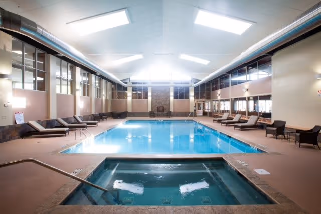 Indoor swimming pool area with a hot tub in the foreground, surrounded by lounge chairs and seating along the sides. The room has large windows, high ceilings with exposed ductwork, and bright overhead lighting.