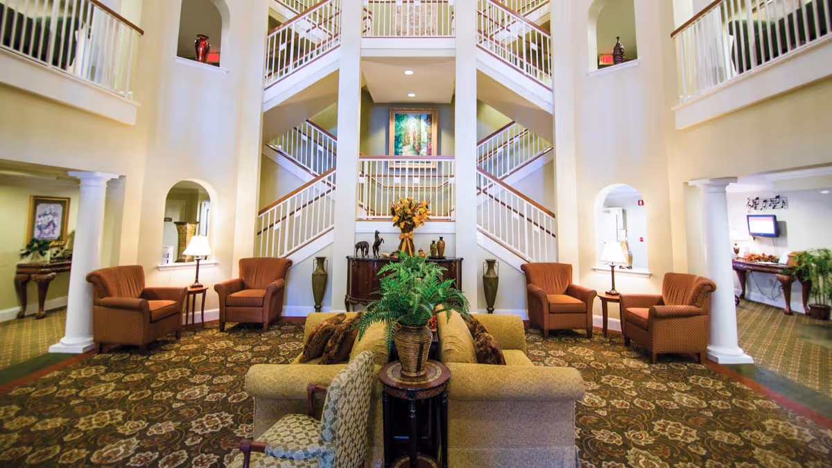 A spacious and well-lit living room area in a senior living facility featuring a central beige sofa with patterned cushions, two matching armchairs on each side, small side tables with lamps, a patterned carpet, decorative plants, and a staircase with white railings in the background.