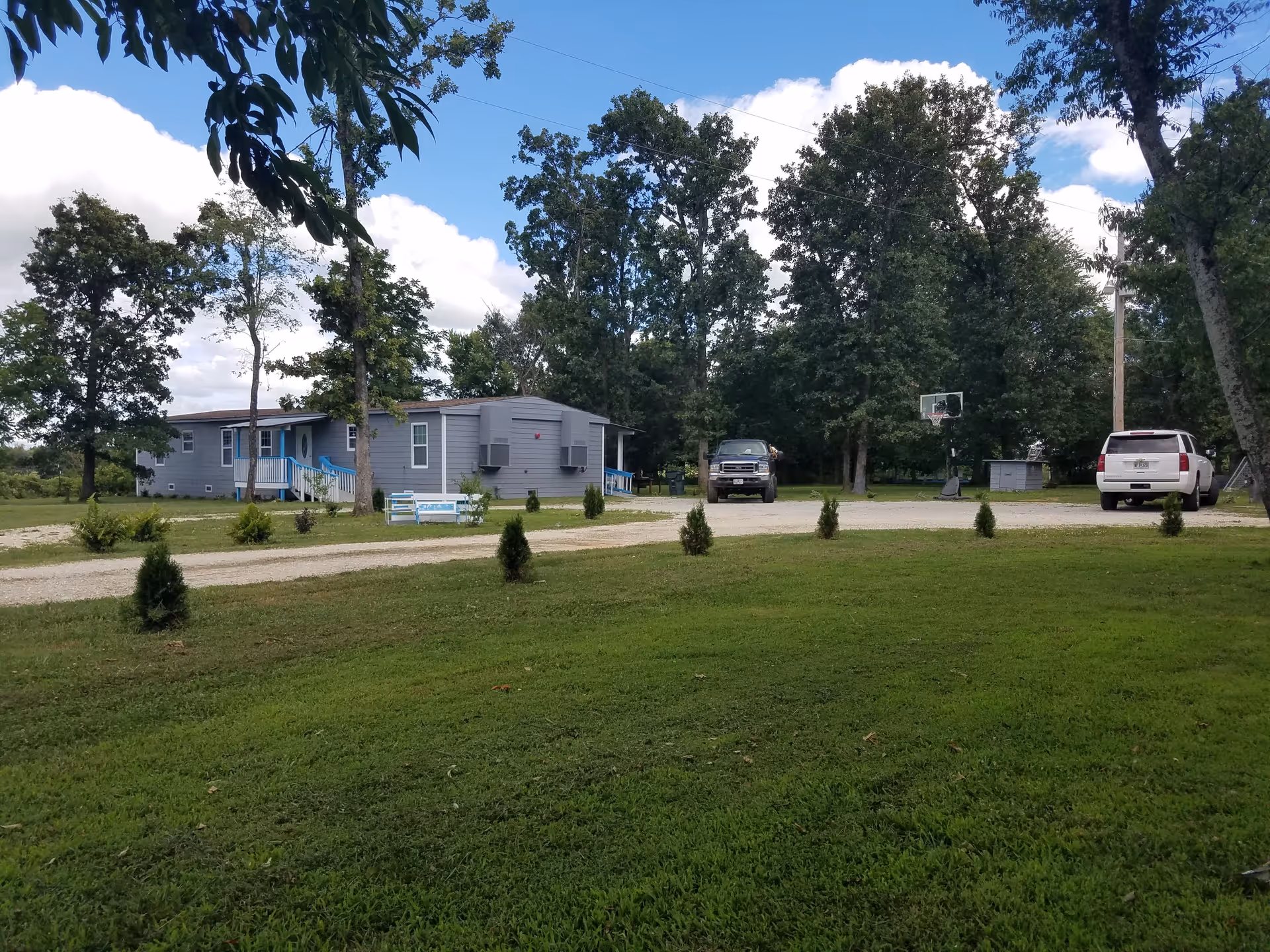 A single-story gray building with blue trim surrounded by trees and grass under a partly cloudy sky. There are two vehicles parked on a gravel driveway, a basketball hoop, and small shrubs planted along the edge of the lawn.