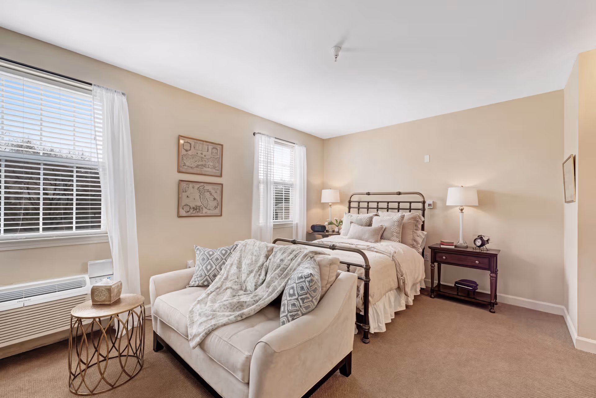 A cozy bedroom with beige walls and carpet, featuring a metal-framed bed with multiple pillows and a beige comforter. In front of the bed is a small beige loveseat with patterned pillows and a throw blanket. There are two windows with white blinds and sheer curtains, a small round side table with a decorative item, and two bedside tables with lamps and an alarm clock.