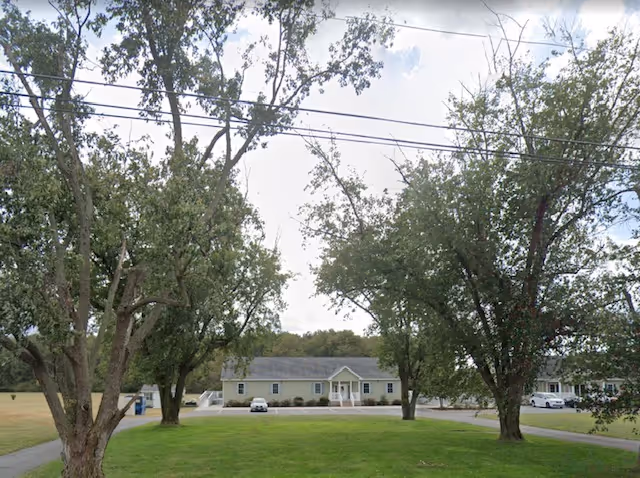 View of a single-story building with a gray roof and beige siding, surrounded by trees and a grassy lawn in the foreground. There are a few cars parked near the building under a partly cloudy sky.