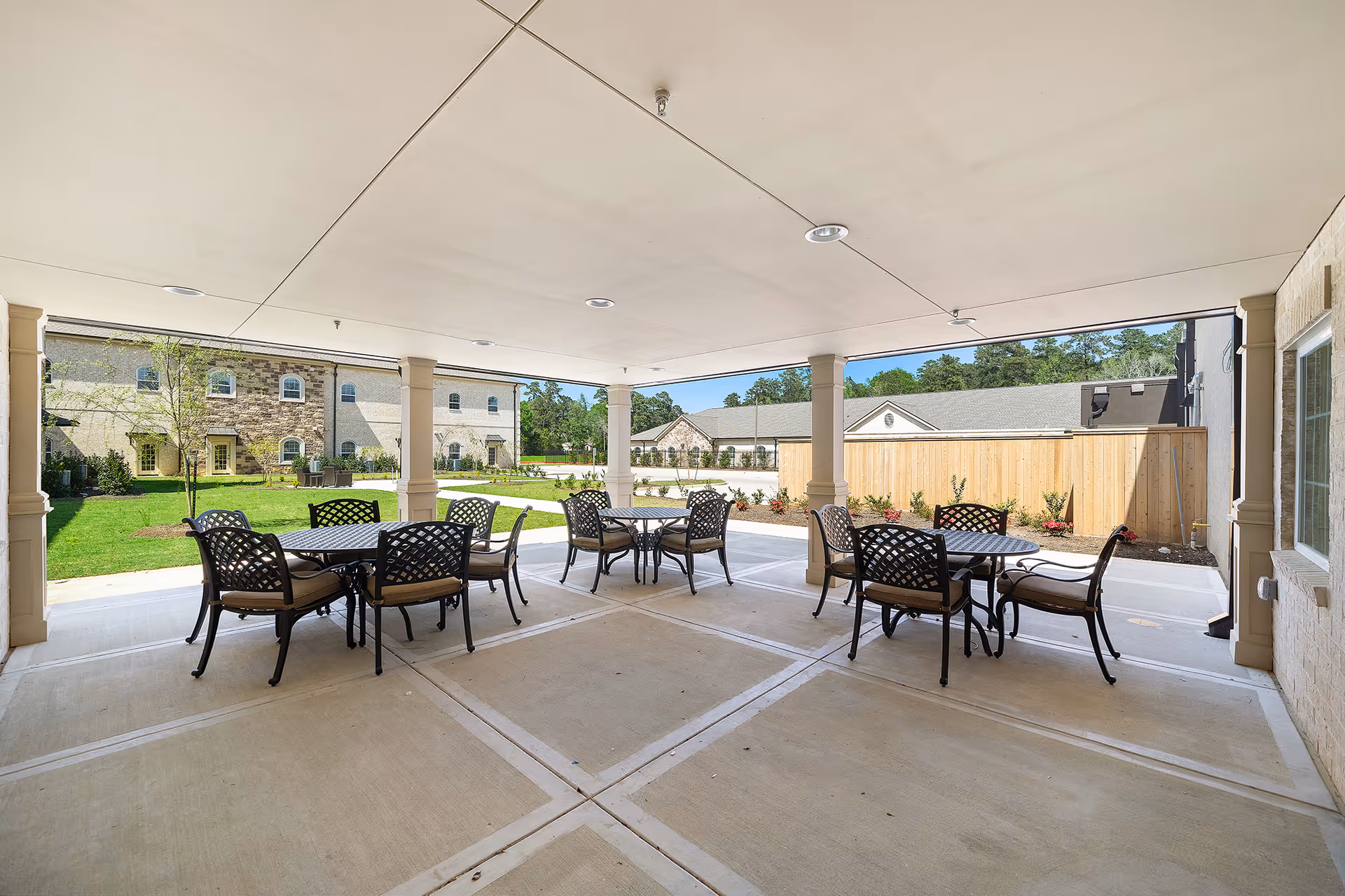 Covered outdoor patio area with several round tables and cushioned chairs arranged for seating. The patio overlooks a green lawn and buildings with stone and beige exteriors under a clear blue sky.